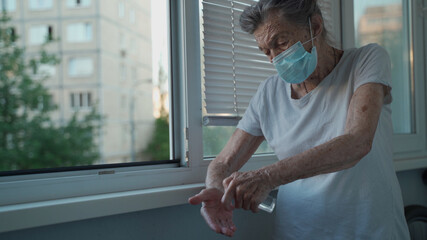 Old woman 90 years wearing face mask use alcohol spray protect covid 19 standing by window in...