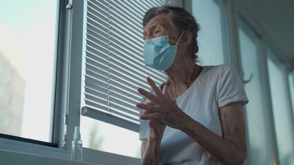 Senior woman applying hand sanitizer, wearing protection mask against Covid-19 standing by the window. Lonely sad older female in face medical mask using hand antibacterial liquid sanitizer at home