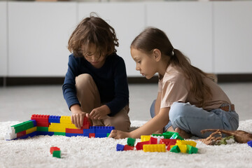 Focused little boy and girl playing construction toys, completing model together, training spatial ability skills. Kids engaged in learning game, enjoying home activity on heating floor with carpet