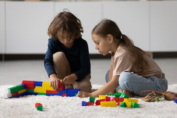 Naklejka premium Focused little boy and girl playing construction toys, completing model together, training spatial ability skills. Kids engaged in learning game, enjoying home activity on heating floor with carpet