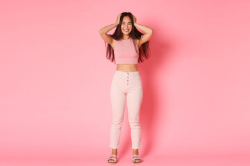 Obraz na płótnie Canvas Fashion, beauty and lifestyle concept. Full length portrait of attractive asian girl in glamour outfit touching hair and smiling broadly, rejoicing over new awesome haircut, pink background