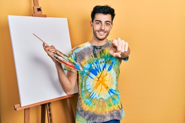 Young hispanic man standing drawing with palette by painter easel stand pointing to you and the camera with fingers, smiling positive and cheerful