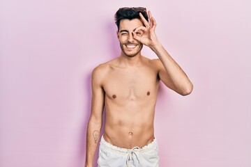 Fototapeta premium Young hispanic man wearing swimwear shirtless doing ok gesture with hand smiling, eye looking through fingers with happy face.