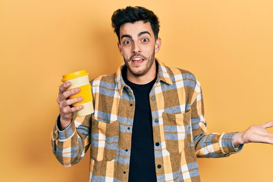 Young hispanic man holding coffee celebrating achievement with happy smile and winner expression with raised hand