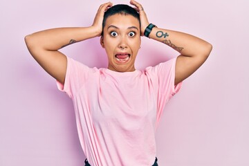 Beautiful hispanic woman with short hair wearing casual pink t shirt crazy and scared with hands on head, afraid and surprised of shock with open mouth