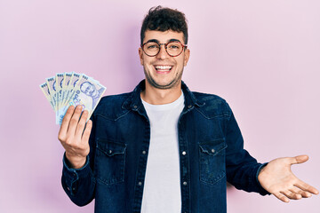 Young hispanic man holding 100 romanian leu banknotes celebrating achievement with happy smile and winner expression with raised hand - obrazy, fototapety, plakaty