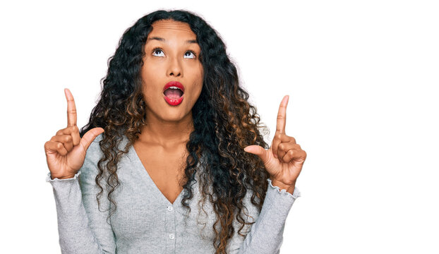 Young hispanic woman with curly hair wearing casual clothes amazed and surprised looking up and pointing with fingers and raised arms.