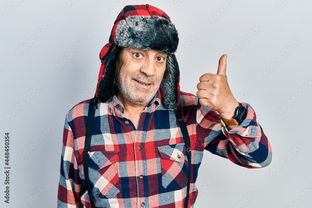 Poster handsome mature man wearing winter hat with ear flaps smiling happy and positive, thumb up doing exc