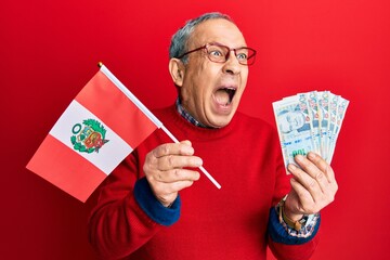 Handsome senior man with grey hair holding peru flag and peruvian sol banknotes angry and mad screaming frustrated and furious, shouting with anger. rage and aggressive concept. - obrazy, fototapety, plakaty