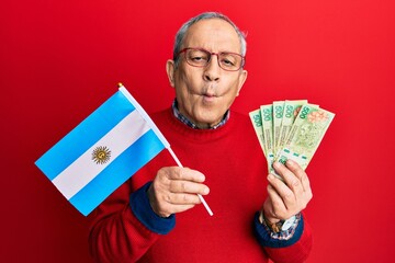 Handsome senior man with grey hair holding argentine flag and pesos banknotes making fish face with...