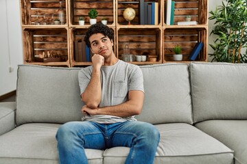 Young hispanic man wearing casual clothes sitting on the sofa at home serious face thinking about...