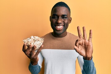 Young african american man holding bowl with pumpkin seeds doing ok sign with fingers, smiling...