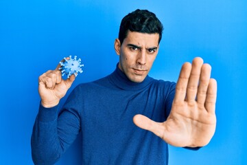 Handsome hispanic man holding virus toy with open hand doing stop sign with serious and confident...