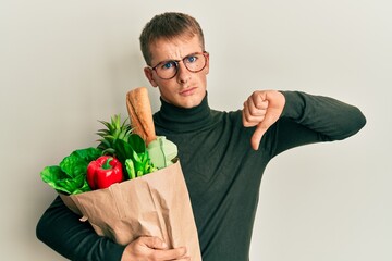 Young caucasian man holding paper bag with groceries with angry face, negative sign showing dislike...