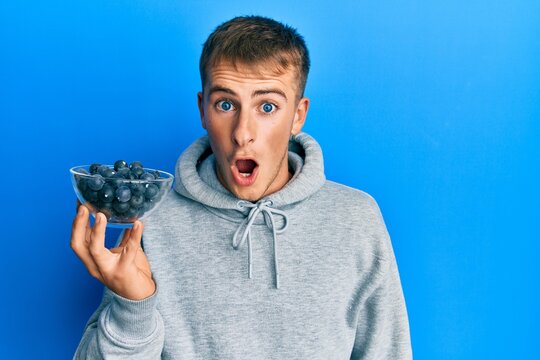 Young caucasian man holding bowl of blueberries scared and amazed with open mouth for surprise, disbelief face