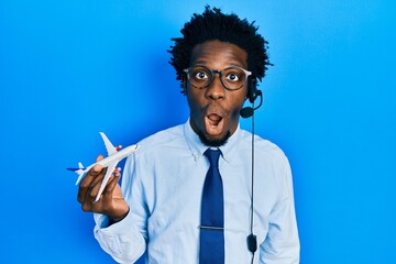 Young african american travel agent man holding plane scared and amazed with open mouth for surprise, disbelief face