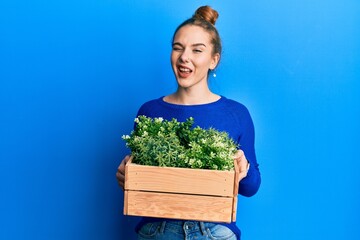Young blonde woman holding wooden plant pot winking looking at the camera with sexy expression,...
