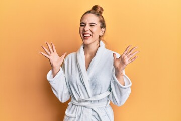Young blonde woman wearing bathrobe celebrating mad and crazy for success with arms raised and...