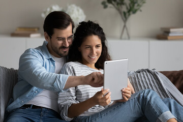 Happy relaxed millennial couple with tablet resting and hugging on couch at home, using online app,...