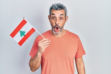 Handsome middle age man with grey hair holding lebanon flag scared and amazed with open mouth for...