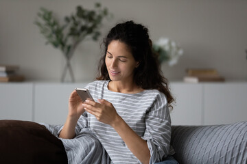 Satisfied mobile phone user receiving message with good news. Happy Latin woman sitting on couch at home, using mobile phone, reading text and smiling, shopping, browsing internet, chatting online