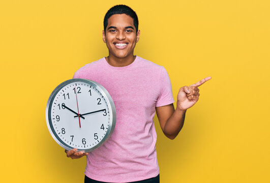 Young handsome hispanic man holding big clock smiling happy pointing with hand and finger to the side