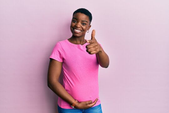 Young african american woman expecting a baby, touching pregnant belly pointing fingers to camera with happy and funny face. good energy and vibes.