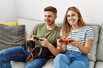 Young hispanic couple smiling happy playing video game sitting on the sofa with dog at home.