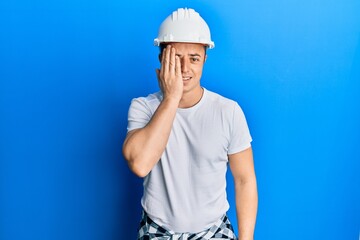 Handsome young man wearing builder uniform and hardhat covering one eye with hand, confident smile on face and surprise emotion.