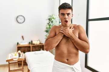 Young hispanic man standing shirtless at spa center shouting and suffocate because painful...