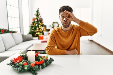 Arab young man sitting on the table by christmas tree doing ok gesture shocked with surprised face, eye looking through fingers. unbelieving expression.