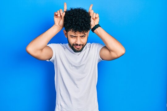 Young arab man with beard wearing casual white t shirt doing funny gesture with finger over head as bull horns