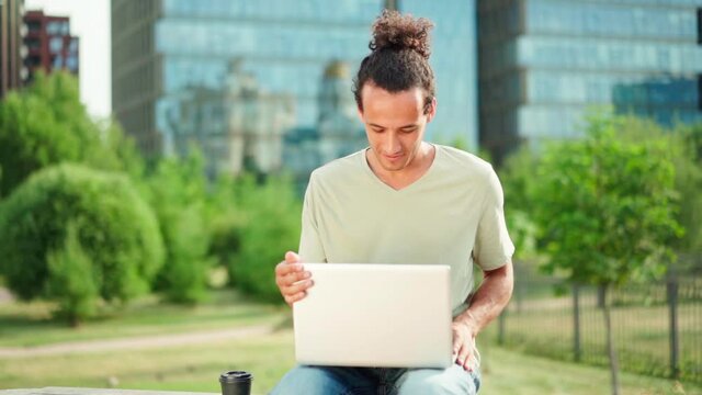 Zoom in slow motion shot of young man working remotely on laptop, closing pc and thinking sitting on bench outdoors. Male entrepreneur, freelancer or student finishing work