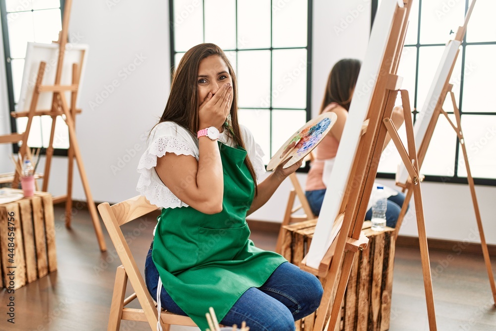 Sticker Young hispanic artist women painting on canvas at art studio laughing and embarrassed giggle covering mouth with hands, gossip and scandal concept