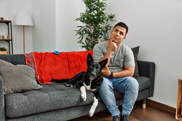 Young latin man and dog sitting on the sofa at home with hand on chin thinking about question, pensive expression. smiling with thoughtful face. doubt concept.