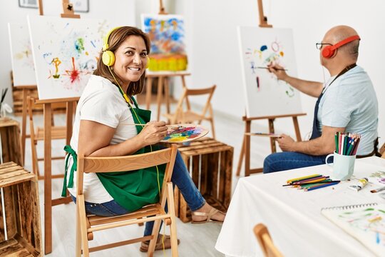 Middle age hispanic painter couple smiling happy using headphones and painting at art studio.