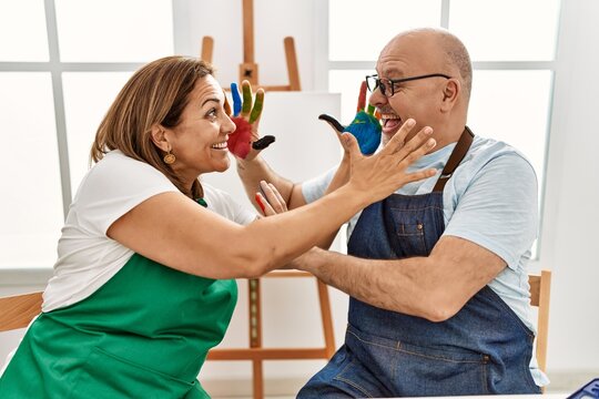 Middle age hispanic painter couple smiling happy touching face with painted hands at art studio.