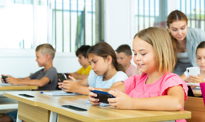 Interested smiling preteen girl using smartphone while studying in schoolroom with classmates and female teacher