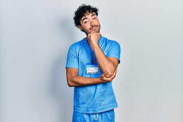Young hispanic man wearing blue male nurse uniform looking confident at the camera with smile with crossed arms and hand raised on chin. thinking positive.