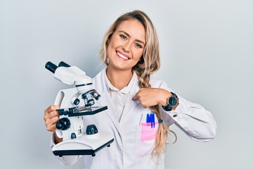 Beautiful young blonde woman holding microscope pointing finger to one self smiling happy and proud