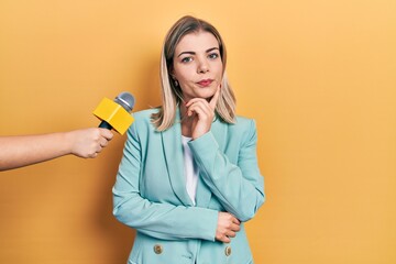 Beautiful caucasian woman being interviewed by reporter holding microphone serious face thinking...