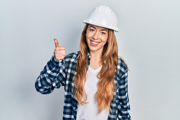 Young caucasian woman wearing architect hardhat smiling happy and positive, thumb up doing excellent and approval sign
