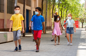 School children in protective medical masks walk along the street of a summer city