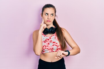 Young brunette teenager wearing gym clothes, using headphones and smart watch serious face thinking...