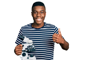 Young african american man holding microscope smiling happy and positive, thumb up doing excellent and approval sign