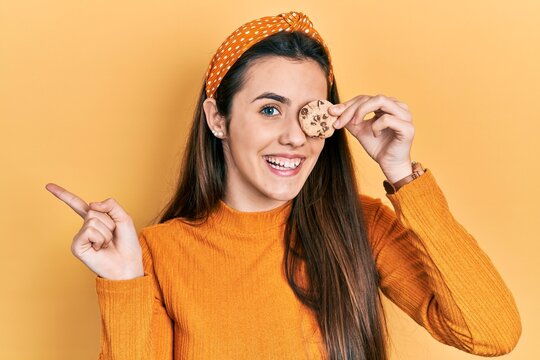 Young brunette teenager holding cookie over eye smiling happy pointing with hand and finger to the side