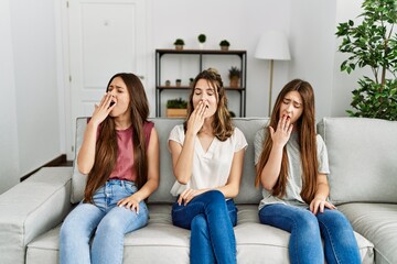 Group of three hispanic girls sitting on the sofa at home bored yawning tired covering mouth with hand. restless and sleepiness.