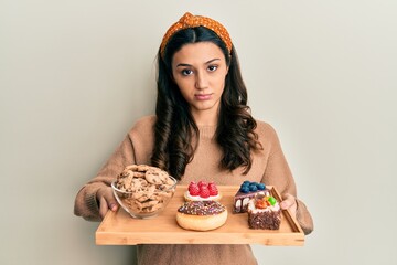 Young hispanic woman holding tray with pastries breakfast skeptic and nervous, frowning upset...