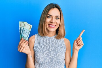 Young caucasian blonde woman holding bangladesh taka banknotes smiling happy pointing with hand and finger to the side