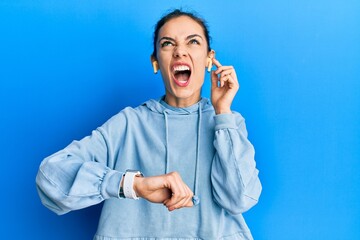 Young caucasian blonde woman using earphones and smart watch angry and mad screaming frustrated and furious, shouting with anger looking up.